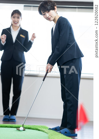 A female president practicing putting indoors and a young employee flattering her A female president practicing putting indoors and a young employee flattering her 122620082