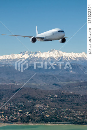 A white wide body passenger airplane flies over resort mountain town on the seashore 122622294