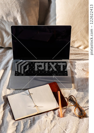 Open laptop, blank notebook and eyeglasses on cozy bed. Minimalist workspace vibe ideal for productivity. 122622413