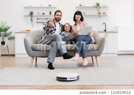 Caucasian family with parents and daughter relaxing on sofa in modern living room with robotic vacuum. Father, mother, and child showing thumbs up. Home technology and bonding scene. 122624632