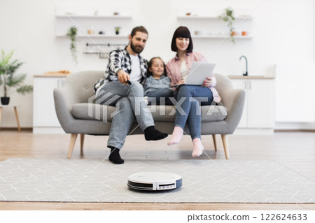 Caucasian family relaxing on sofa with robotic vacuum cleaning floor. Young parents and daughter enjoying technology at home. Modern lifestyle with white man, woman, and girl, aged 30s and 10s. 122624633