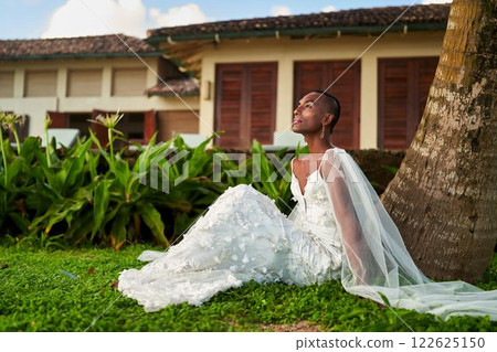 Gender fluid person in white wedding dress sits by palm tree, embracing tropical scenery. LGBTQ bridal concept. Calm moment at exotic destination. Modern identity, style in inclusive wedding fashion. Gender fluid person in white wedding dress sits by palm tree, embracing tropical scenery. LGBTQ bridal concept. Calm moment at exotic destination. Modern identity, style in inclusive wedding fashion. 122625150