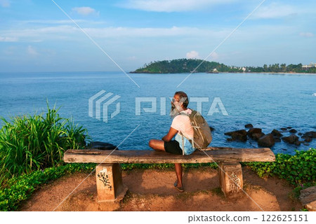 Bearded senior man with backpack relaxes on bench by ocean. He contemplates nature and life. Scene suggests travel, reflection. Ideal for themes of introspection, healing, mental health, loneliness. Bearded senior man with backpack relaxes on bench by ocean. He contemplates nature and life. Scene suggests travel, reflection. Ideal for themes of introspection, healing, mental health, loneliness. 122625151