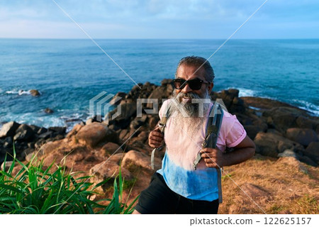 Senior ethnic man hikes on rocky coastal path, enjoys nature. Ocean view, backpack adventure. Elderly active lifestyle, explore outdoors. Sunny day, rugged terrain, vibrant sea backdrop. 122625157