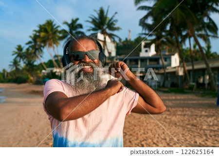 Elderly gray-bearded man dances on tropical beach with wireless headphones. Stylish senior enjoys sunny day and music. Trendy scene for summer vibes, vacation energy, beach party. 122625164