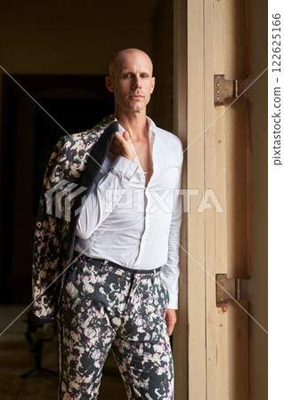 Bald man in striking floral suit in luxury villa. Bold gay groom outfit, confident. Elegant fashion statement at villa wedding. Unique attire, style innovation. Modern LGBTQ fashion. 122625166