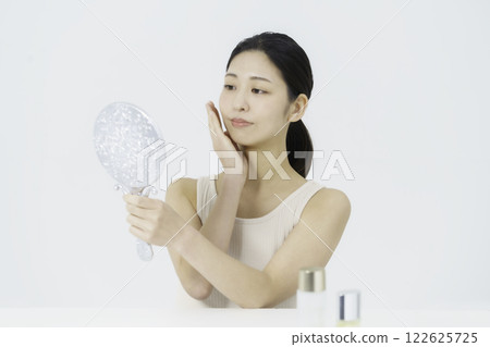 A smiling young woman wearing a tank top on a white background and holding her hand on her cheek while looking in the mirror Beauty A smiling young woman wearing a tank top on a white background and holding her hand on her cheek while looking in the mirror Beauty 122625725