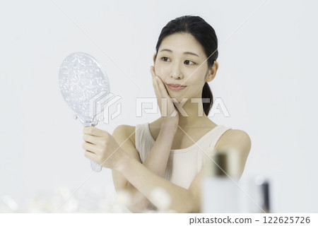A smiling young woman wearing a tank top on a white background and holding her hand on her cheek while looking in the mirror Beauty A smiling young woman wearing a tank top on a white background and holding her hand on her cheek while looking in the mirror Beauty 122625726