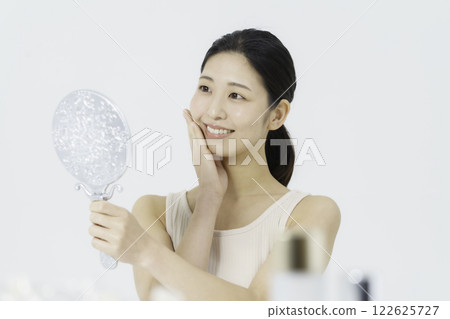 A smiling young woman wearing a tank top on a white background and holding her hand on her cheek while looking in the mirror Beauty 122625727