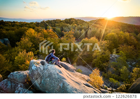 Aerial view of climber sitting atop rock formation at Dovbush Rocks in Carpathian mountains, Ukraine. Sun sets, casting warm glow over autumn forest and distant hills, creating breathtaking view. 122626878