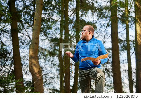Man traveler in glasses stands in dense forest, holding map in one hand and compass in other. Tourist planning outdoor adventure or hiking route, amidst tall trees and natural surroundings. 122626879