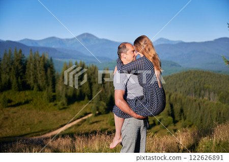 Couple shares intimate moment in scenic mountain setting. Man holds woman close to him as they embrace and look into each other's eyes. Lush green forest and distant mountains under clear blue sky. Couple shares intimate moment in scenic mountain setting. Man holds woman close to him as they embrace and look into each other's eyes. Lush green forest and distant mountains under clear blue sky. 122626891