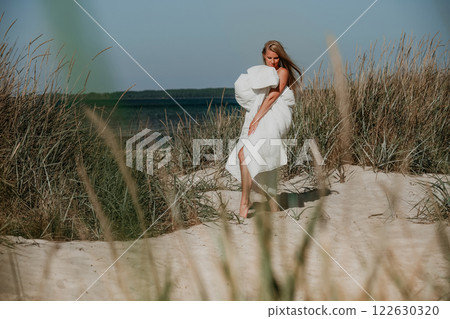 Woman wrapped in white blanket walking barefoot through sandy grasslands near the ocean on sunny day Woman wrapped in white blanket walking barefoot through sandy grasslands near the ocean on sunny day 122630320