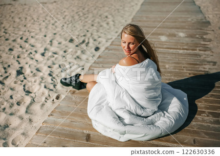 A serene beach moment with a woman wrapped in white blanket enjoying the sun on a wooden dock by the A serene beach moment with a woman wrapped in white blanket enjoying the sun on a wooden dock by the 122630336