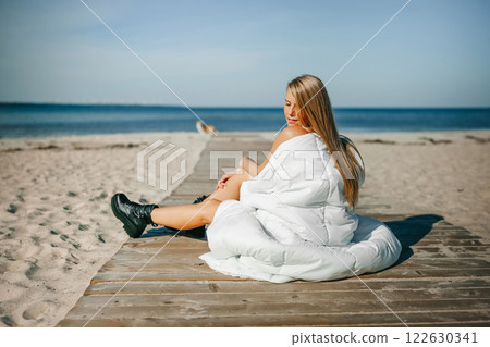 A serene beach moment with a woman wrapped in white blanket enjoying the sun on a wooden dock by the A serene beach moment with a woman wrapped in white blanket enjoying the sun on a wooden dock by the 122630341