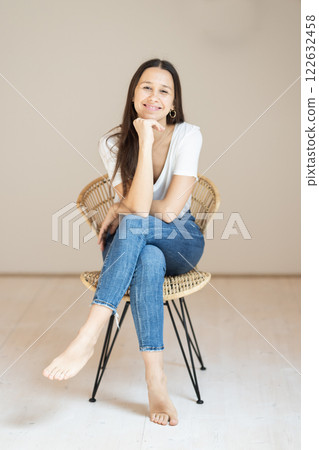 Portrait of confident beautiful woman with long brown hair, wearing casual clothes, sitting on chair in tight jeans and white t-shirt, studio background Portrait of confident beautiful woman with long brown hair, wearing casual clothes, sitting on chair in tight jeans and white t-shirt, studio background 122632458