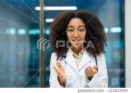 African American woman doctor wearing white coat in modern hospital, engaged in video call. Confident expression, looking at camera, conveying care, professionalism. Ideal for medical, technology 122632485