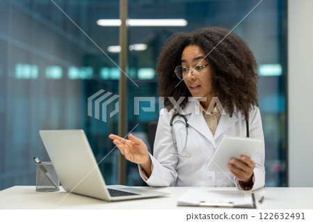 African American woman doctor engaging in online consultation using laptop and tablet in office. Professional healthcare, technology, patient communication, and expertise conveyed. 122632491