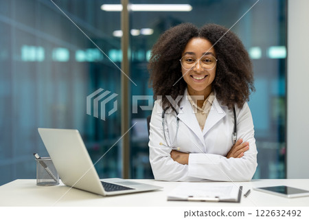 Confident African American female doctor in white coat with smile arms crossed in modern office. Afro hair, glasses on, with laptop, clipboard. Professional medical setting, healthcare concept. 122632492