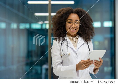African American woman doctor wearing white coat and stethoscope, smiling while holding tablet. Represents professionalism, healthcare, technology in modern medical environment. 122632592
