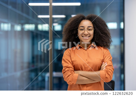 Confident young professional woman with glasses standing arms crossed, smiling warmly in modern office. Represents empowerment, success, and leadership in business setting. Confident young professional woman with glasses standing arms crossed, smiling warmly in modern office. Represents empowerment, success, and leadership in business setting. 122632620