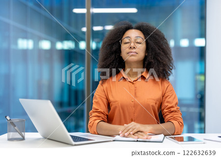 Calm and focused woman breathing and resting relaxing sitting at desk inside office. Young businesswoman working with laptop. 122632639