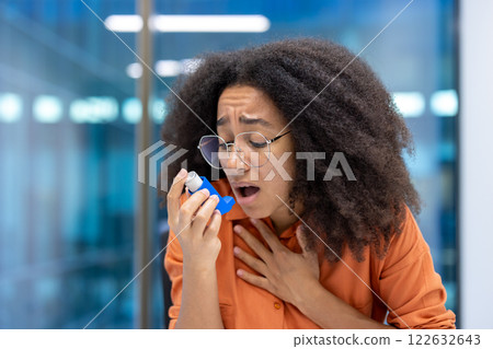 Concerned young woman using asthma inhaler in modern office. She holds her chest, appearing distressed. Office setting visible in background, conveying need for health care support. Concerned young woman using asthma inhaler in modern office. She holds her chest, appearing distressed. Office setting visible in background, conveying need for health care support. 122632643