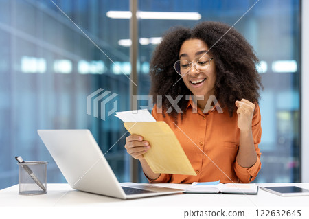 Joyful woman in orange shirt celebrates good news from letter at office desk. She smiles, fist raised, beside laptop. Concepts include success, happiness, achievement, communication. Joyful woman in orange shirt celebrates good news from letter at office desk. She smiles, fist raised, beside laptop. Concepts include success, happiness, achievement, communication. 122632645