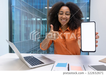 Smiling woman with glasses and afro hair holds smartphone with blank screen and gives thumbs-up gesture, seated at desk with laptop in office. Ideal for concepts of positivity, technology, approval. 122632671