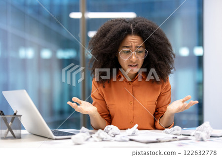 Frustrated woman in office surrounded by crumpled papers, laptop open, expressing confusion. Business setting illustrates work challenge, stress. Represents problem-solving, stress, frustration. 122632778
