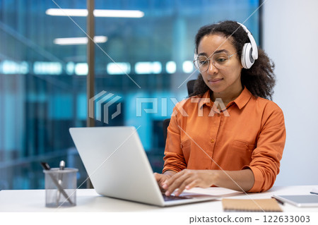 Young African American businesswoman working on laptop in modern office setting. Wearing headphones, she appears focused and engaged. Ideal for themes of technology, business, and productivity. Young African American businesswoman working on laptop in modern office setting. Wearing headphones, she appears focused and engaged. Ideal for themes of technology, business, and productivity. 122633003