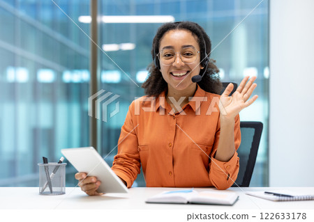 Confident African American businesswoman in modern office smiling, waving during video call. Wearing headset, holding tablet, radiating positivity and professionalism, showcasing remote communication Confident African American businesswoman in modern office smiling, waving during video call. Wearing headset, holding tablet, radiating positivity and professionalism, showcasing remote communication 122633178