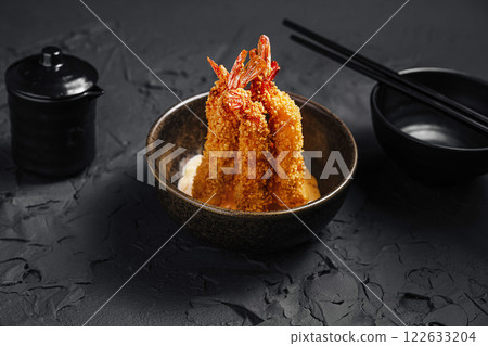 Crispy breaded shrimp served in a dark bowl with condiments and chopsticks on a textured black surface 122633204