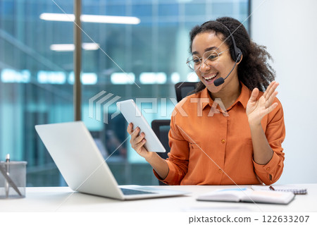 Office worker using smartphone and laptop with headset in modern workspace, engaging in online meeting, multitasking across devices, showcasing professional communication skills and remote work setup 122633207