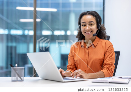 Happy customer service professional wearing headset, working at office desk with laptop, embodying friendly business communication, technology, and support. 122633225