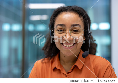Smiling young African American office worker wearing glasses and headset in modern setting shows confidence in customer support. Represents professionalism and positive attitude in workplace. 122633226