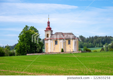 The Church of Saint John the Baptist stands serenely in Vidonice, Czechia, surrounded by lush green fields and trees, reflecting a tranquil rural scene under a clear blue sky. 122633258