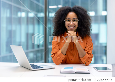 Confident woman in office setting with laptop and open book. She exudes professionalism, approachability, wearing glasses and orange blouse, symbolizing perfect blend of work and positivity. 122633627