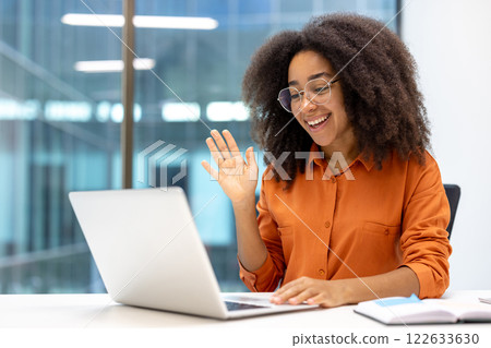 Smiling woman in orange shirt waves during online meeting, engaging in communication through laptop. Bright office setting conveys positive work-from-home environment and internet connectivity. Smiling woman in orange shirt waves during online meeting, engaging in communication through laptop. Bright office setting conveys positive work-from-home environment and internet connectivity. 122633630