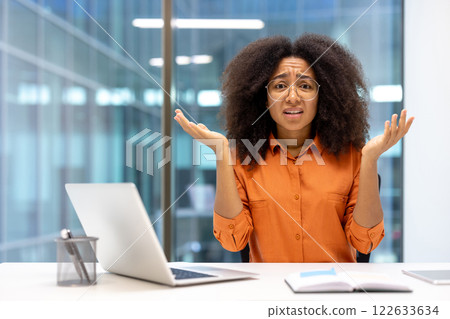 Confused woman in office setting expressing uncertainty. Dressed in orange shirt, sitting at desk with open laptop and book, raising hands in confusion, seeking help. 122633634