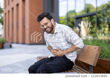 Contorted from severe pain, a young Indian man sits on a bench in the street and holds his stomach with his hands. Contorted from severe pain, a young Indian man sits on a bench in the street and holds his stomach with his hands. 122633682