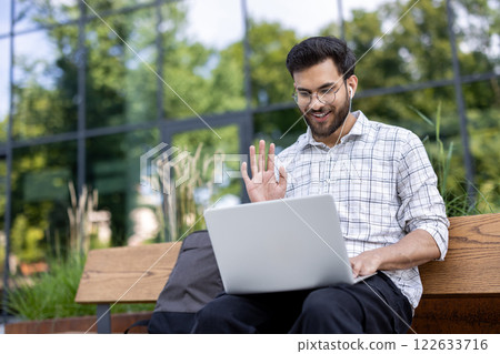 Smiling man using laptop outdoors holding video call with headphones wearing casual attire. Background of modern building , emphasizing connection, technology, and remote communication Smiling man using laptop outdoors holding video call with headphones wearing casual attire. Background of modern building , emphasizing connection, technology, and remote communication 122633716