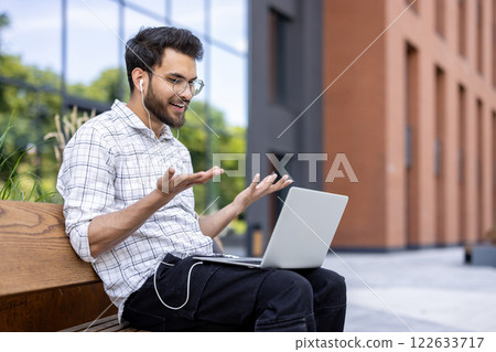 Smiling Indian young man sitting on a bench outside an office center wearing headphones and talking on a video call ,on a laptop while gesturing with his hands. 122633717