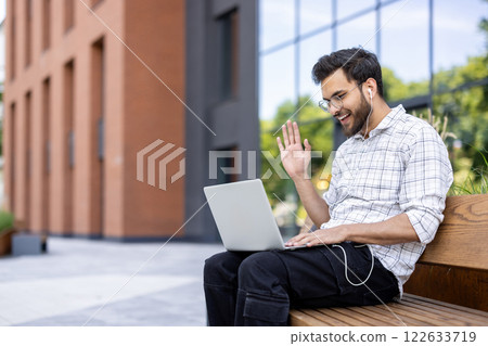 Man using laptop for video call sitting on bench outside office building wearing earphones. Person smiling and waving, engaging in online communication with phone 122633719