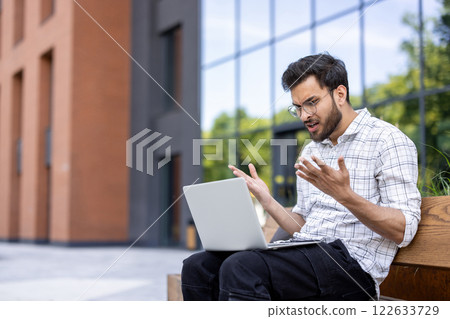 Young man shows frustration while working on laptop outdoors. Urban setting with modern office buildings in background. Difficulties with technology, work stress, and problem solving portrayed 122633729