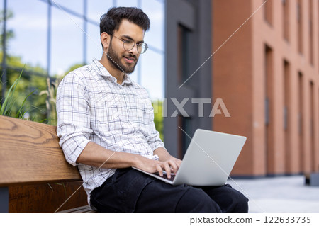 Young man sitting on bench using laptop. Outdoor setting near modern building, focusing on work or study. Warm and productive atmosphere. Young man sitting on bench using laptop. Outdoor setting near modern building, focusing on work or study. Warm and productive atmosphere. 122633735