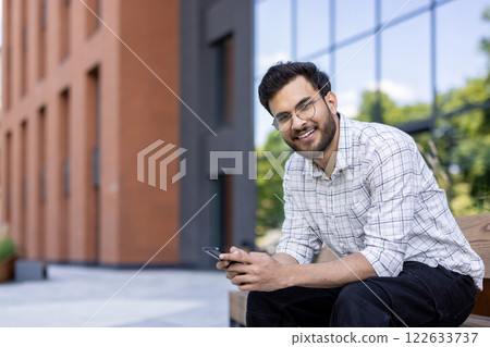 Man wearing glasses sitting on bench outside modern building holding smartphone. Smiling relaxed individual enjoying outdoor environment with blurred urban architecture background. C Man wearing glasses sitting on bench outside modern building holding smartphone. Smiling relaxed individual enjoying outdoor environment with blurred urban architecture background. C 122633737