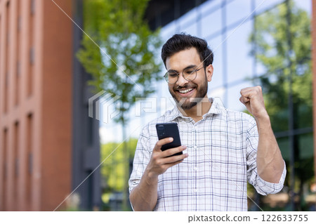 Young man wearing glasses and plaid shirt celebrating success while looking at phone outdoors. Cheerful man with clenched fist standing near building and glass windows, expressing joy 122633755