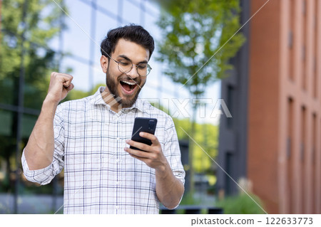 Photo of a happy young Indian man standing outside on the street, looking at the phone screen and rejoicing in success, raising his hand up. Photo of a happy young Indian man standing outside on the street, looking at the phone screen and rejoicing in success, raising his hand up. 122633773