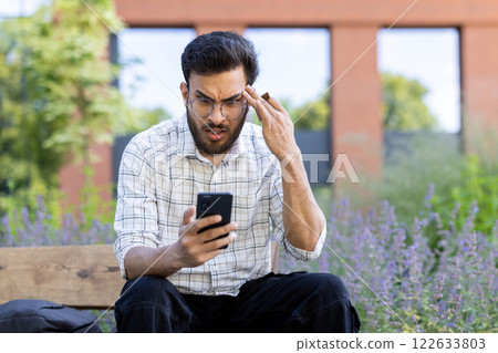 Man sitting on bench in park checking smartphone with worried expression. Urban area in background with greenery. Concept of stress, technology, communication. 122633803
